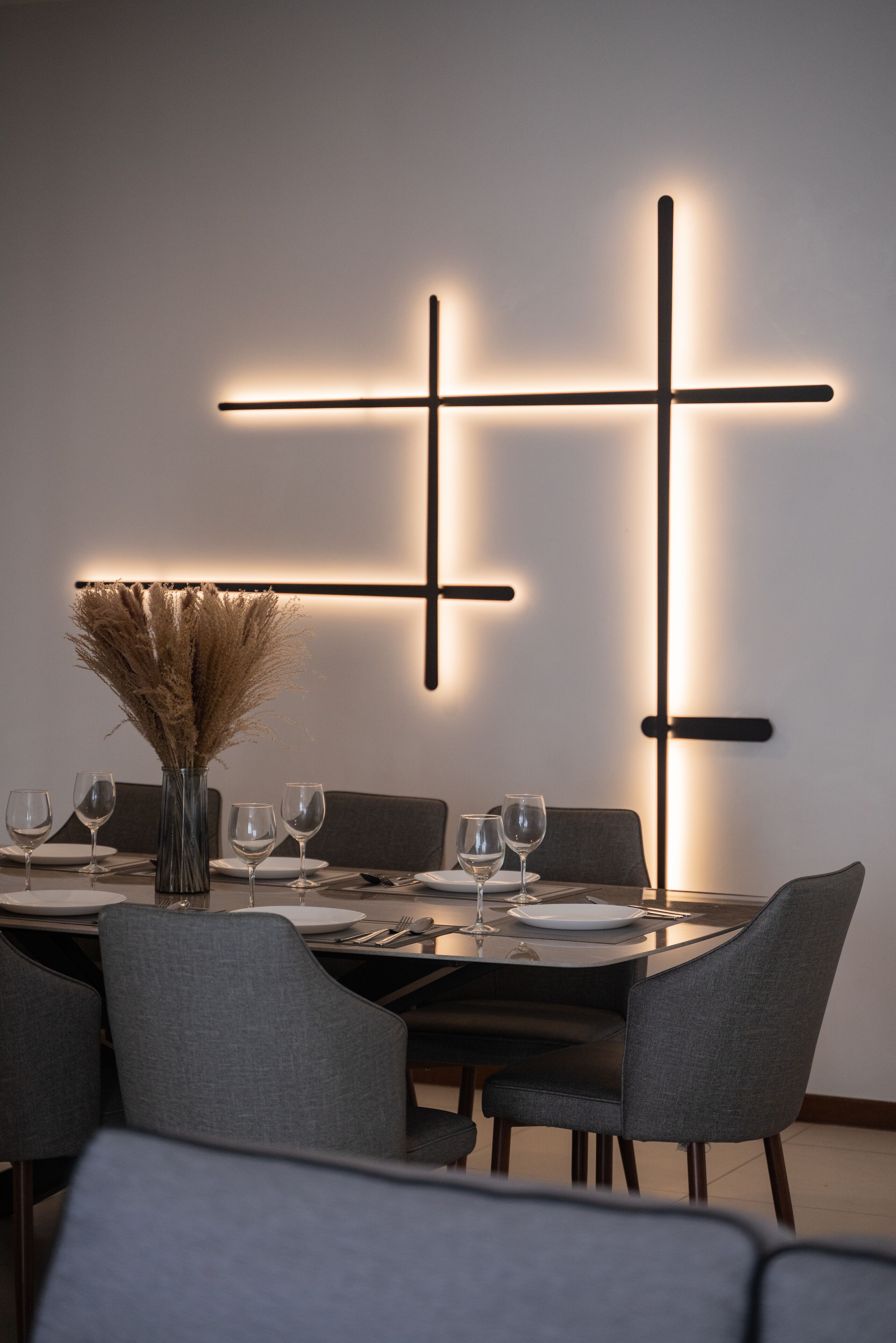 A modern dining area with a minimalist table set for four, featuring a vase of dried plants, a glass of water, and a wine glass.