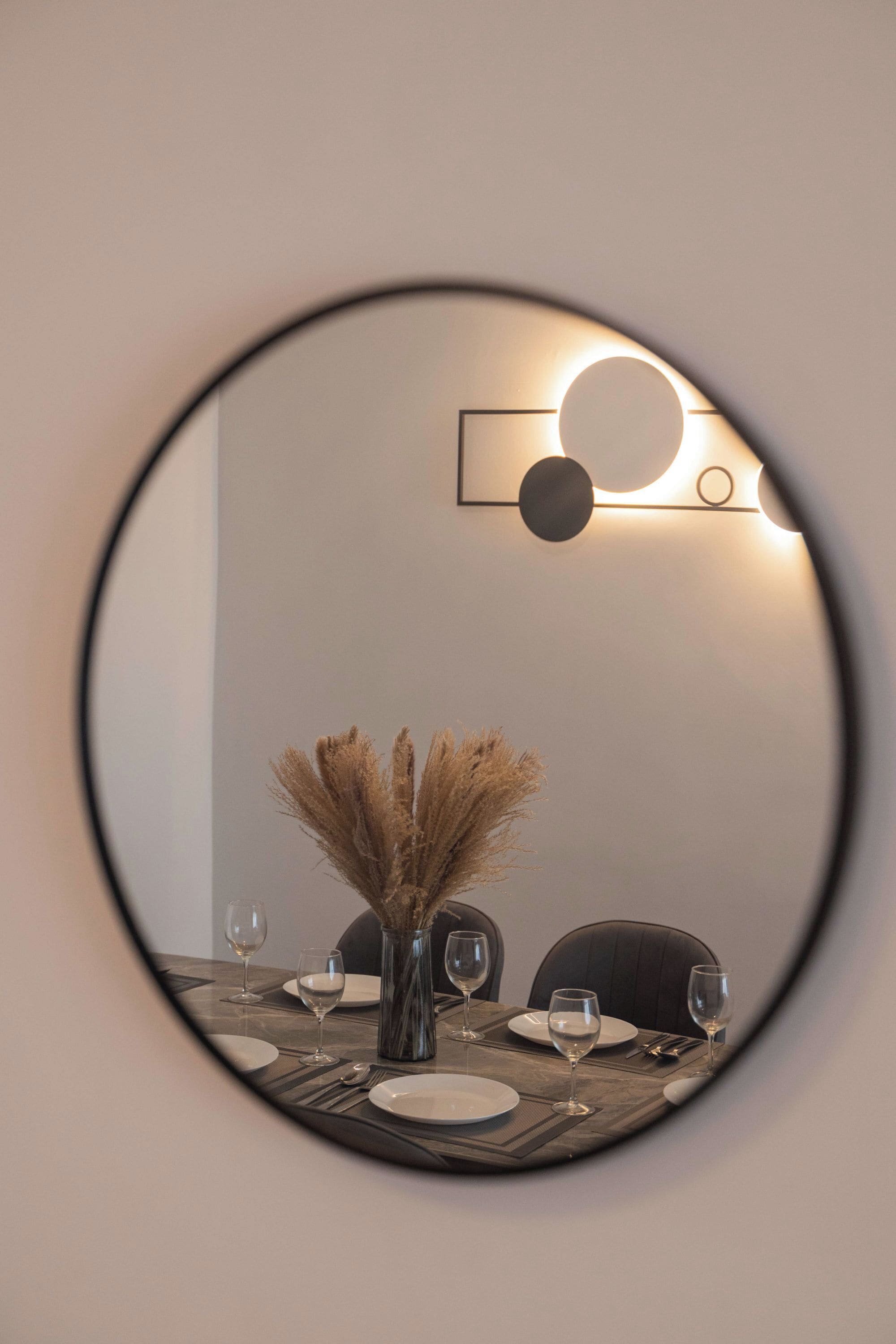 A round mirror reflecting a dining table with a vase of dried plants, wine glasses, and cutlery.