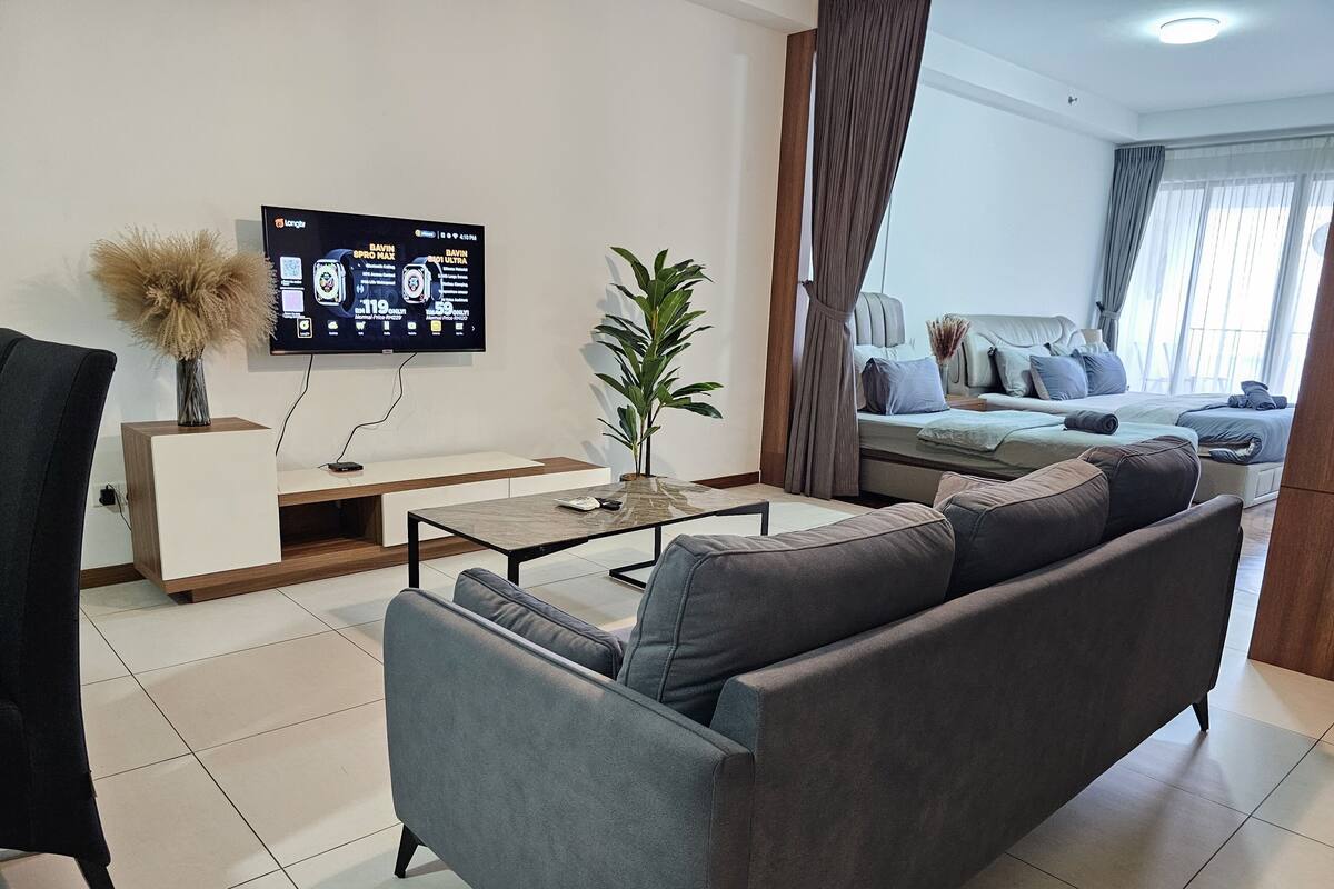 Ein modernes Wohnzimmer mit grauem Sofa, Couchtisch, Wand-Fernseher, Topfpflanze und Bett mit grauem Kopfteil.