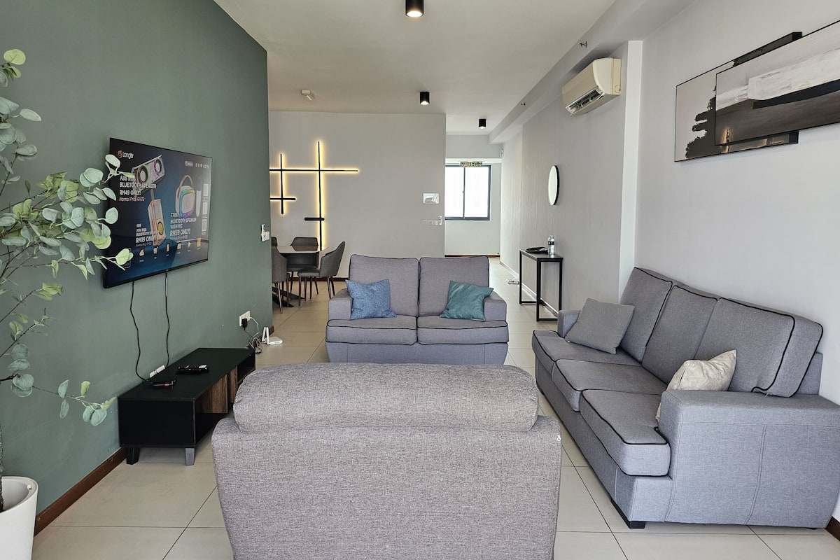 Ein modernes Wohnzimmer mit einem grauen Sofa, einem Couchtisch und einem an der Wand montierten Fernseher. Im Hintergrund befindet sich ein Essbereich mit Tisch und Stühlen.