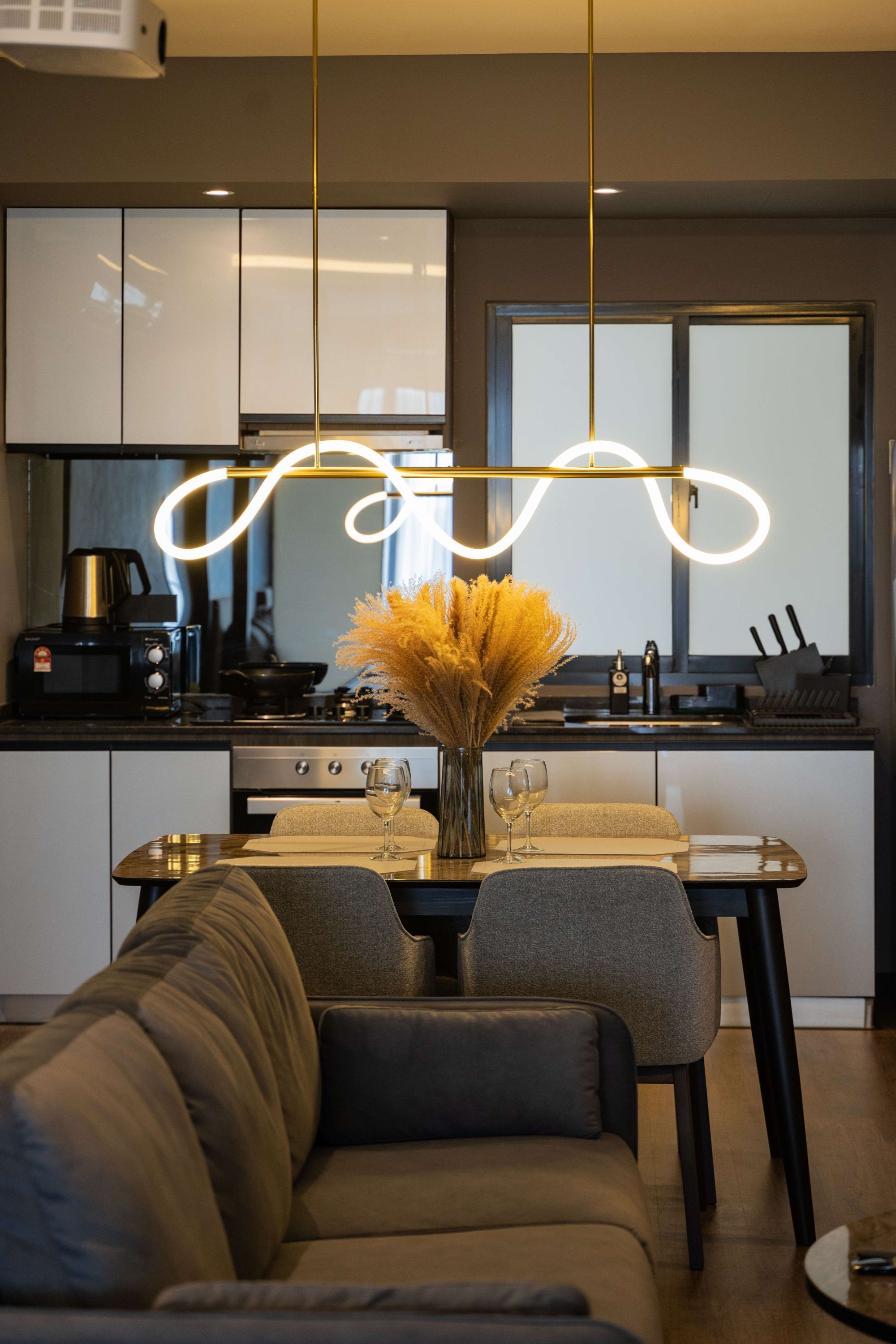 A modern kitchen with a dining area, featuring a sofa, a table with wine glasses, and a vase with dried flowers.