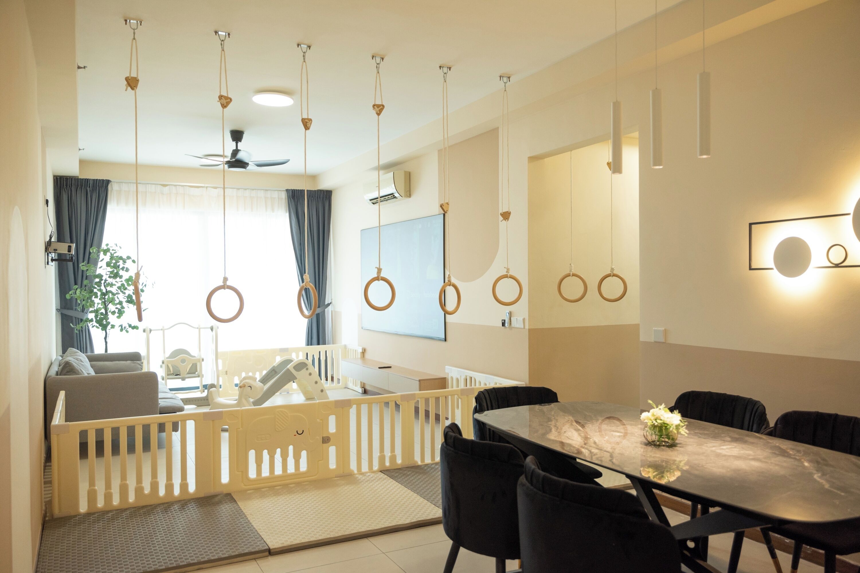 A modern living room with a dining area, featuring a high ceiling, pendant lights, a ceiling fan, and a large window with curtains.