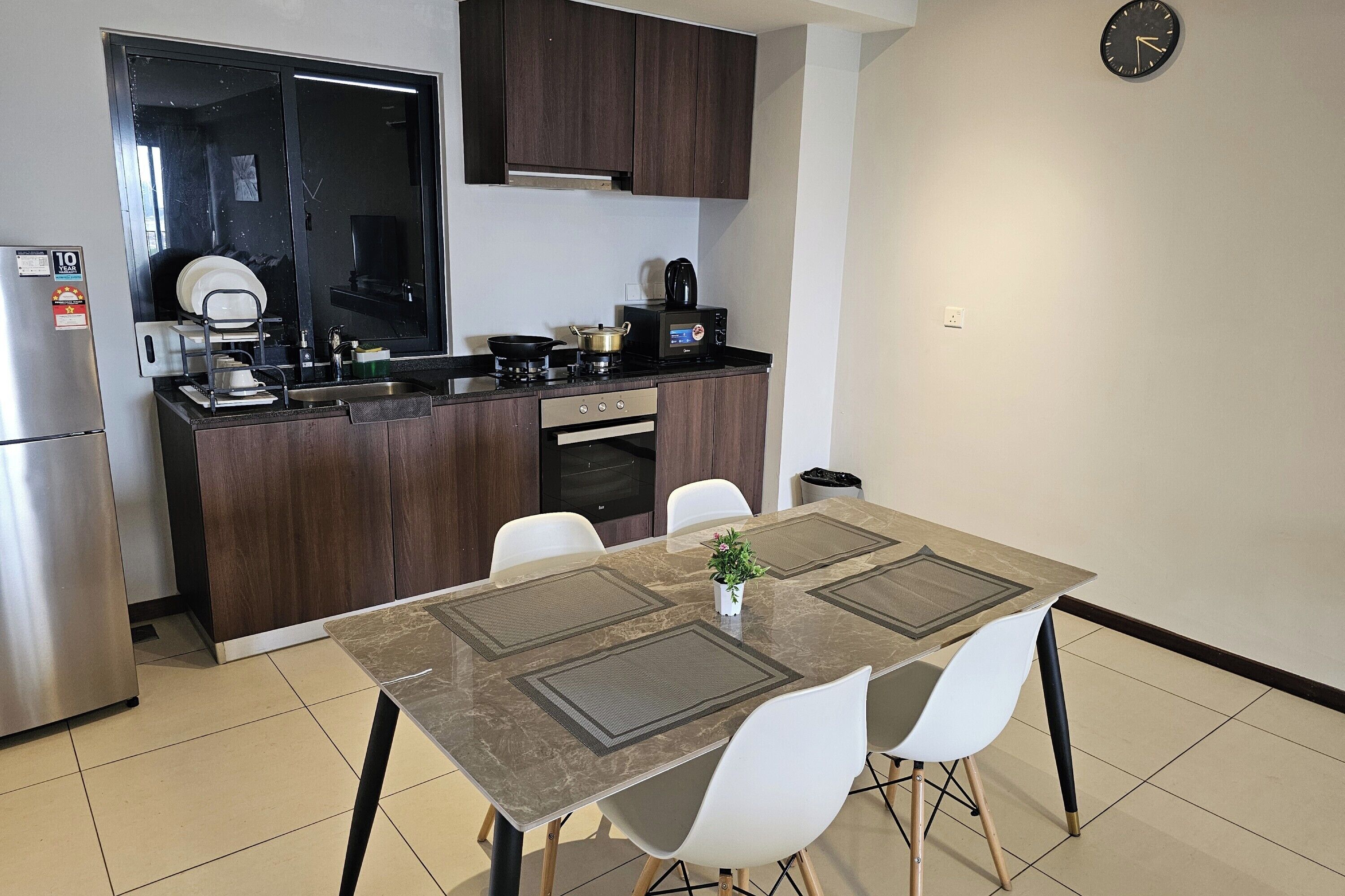 A compact kitchen with dark wood cabinets, a stainless steel refrigerator, a microwave, and a stove. There is a dining table with four white chairs and a small potted plant.