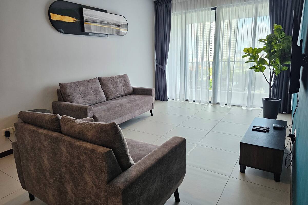 Ein modernes Wohnzimmer mit einem grauen Sofa, einem kleineren Sessel, einem Beistelltisch mit Pflanze und einem großen Fenster mit transparenten Vorhängen.