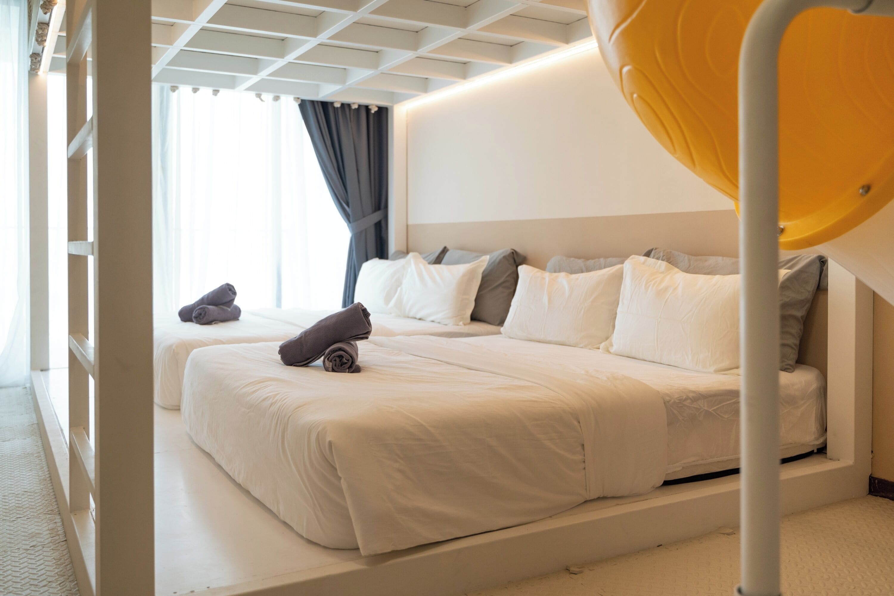 Una habitació d'hotel amb dos llits, un gran finestral amb cortines i una làmpada de sostre de color groc peculiar.