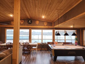 Un interior de madera con una mesa de billar, sillas y grandes ventanales con vistas a un lago.