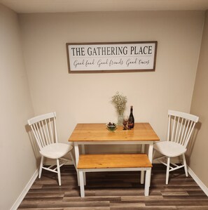 A dining area with a wooden table and two white chairs, a sign above stating "THE GATHERING PLACE", and a vase with flowers on the table.