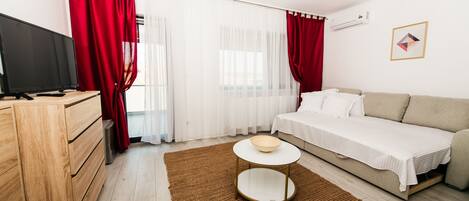 A modern hotel room with a wooden dresser, a television, a bed with a white bedspread, a brown rug, and a small round table with a bowl.