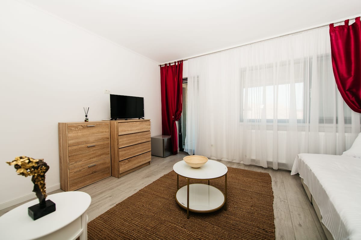A hotel room with a bed, a TV on a wooden cabinet, a small round table with a bowl, and a red curtain.