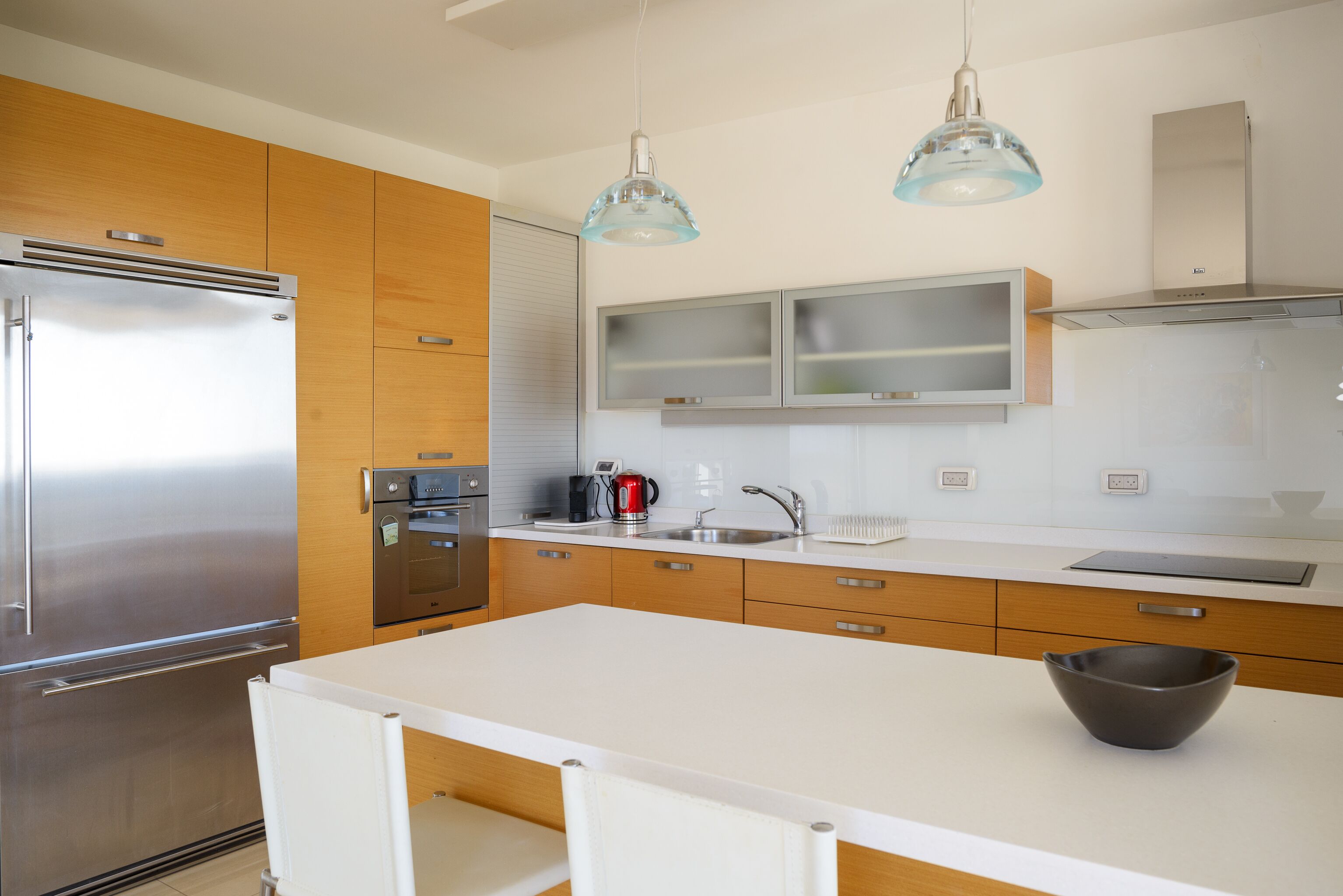 A modern kitchen with a stainless steel refrigerator, wooden cabinets, a white countertop, and two pendant lights.