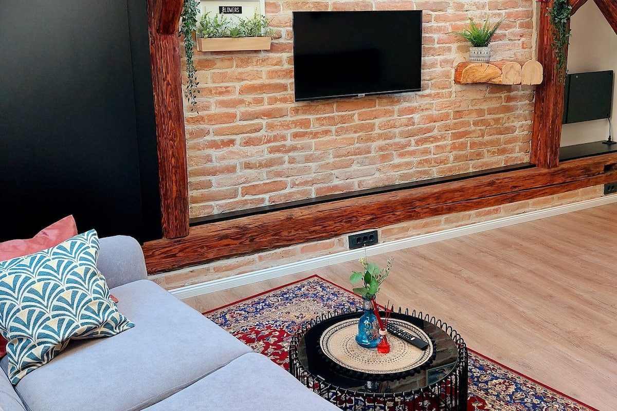 Ein Loft-Wohnzimmer mit freiliegenden Holzbalken, einer Ziegelwand, einem Flachbildfernseher, einer Couch mit gemusterten Kissen und einer Zieruhr.