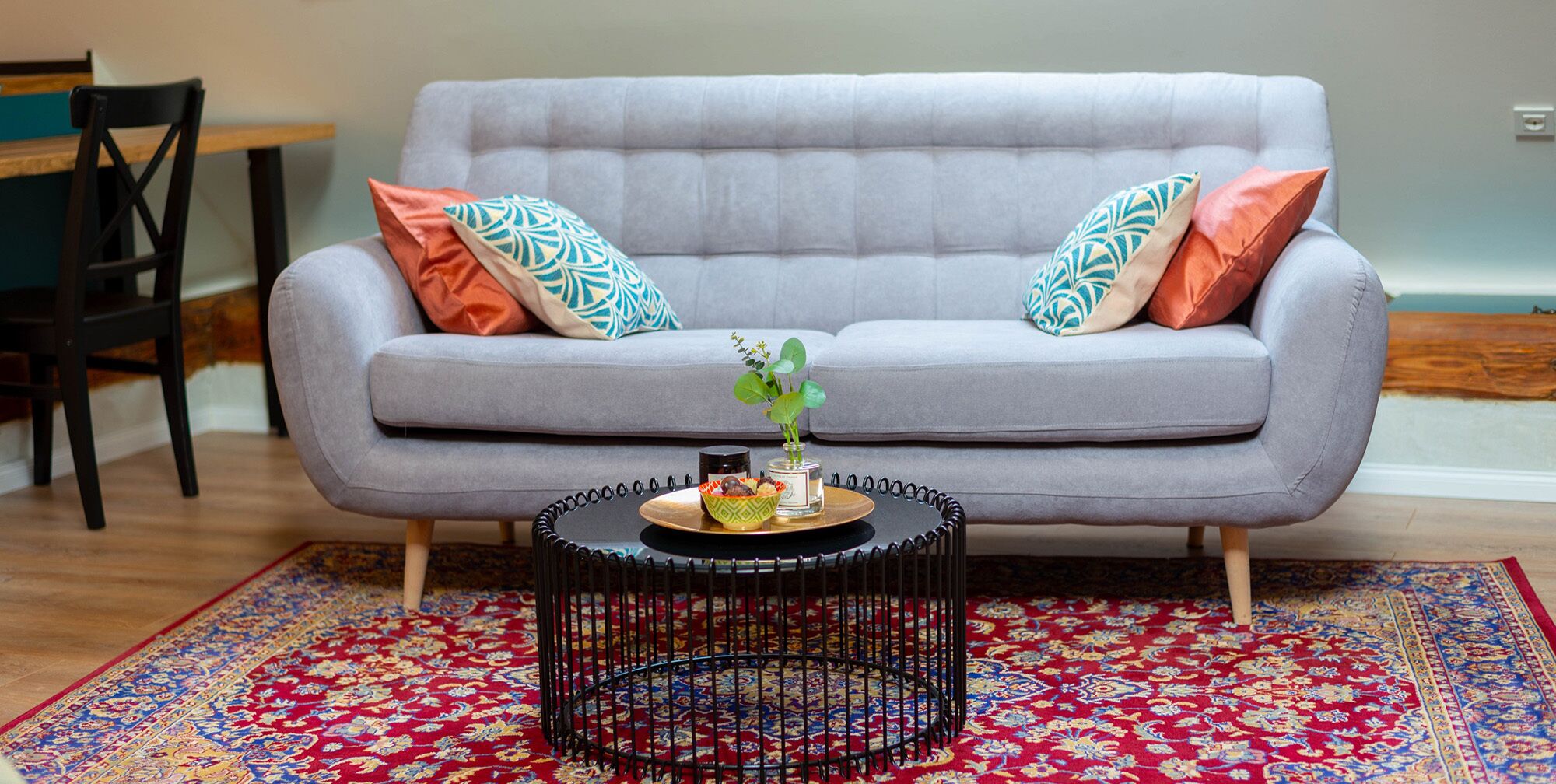 Un canapé gris agrémenté de coussins à motifs, une petite table ronde avec une plante et un bol, et un tapis rouge à motifs.