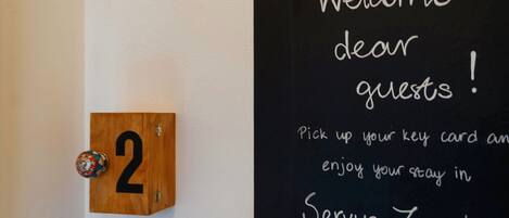 A wooden door with the number 2 and a key card holder. A blackboard with "Welcome dear guests!" and "Pick up your key card and enjoy your stay in Servus Zaagreb" is visible.