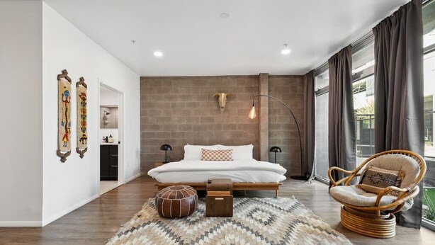 Ein modernes Schlafzimmer mit einem großen Bett, Holzboden, einem gewebten Teppich, einem Korbstuhl und einer Wandleuchte.