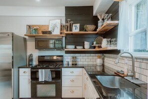 A compact kitchen with a built-in microwave, stove, and sink. There are wooden shelves above the counter holding various kitchen items. - A beautiful home near OSU campus (Corvallis)