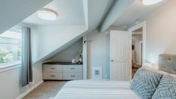 Quarto com teto inclinado, cama com roupa de cama listrada, mesa de cabeceira, uma cadeira e janela com cortinas.