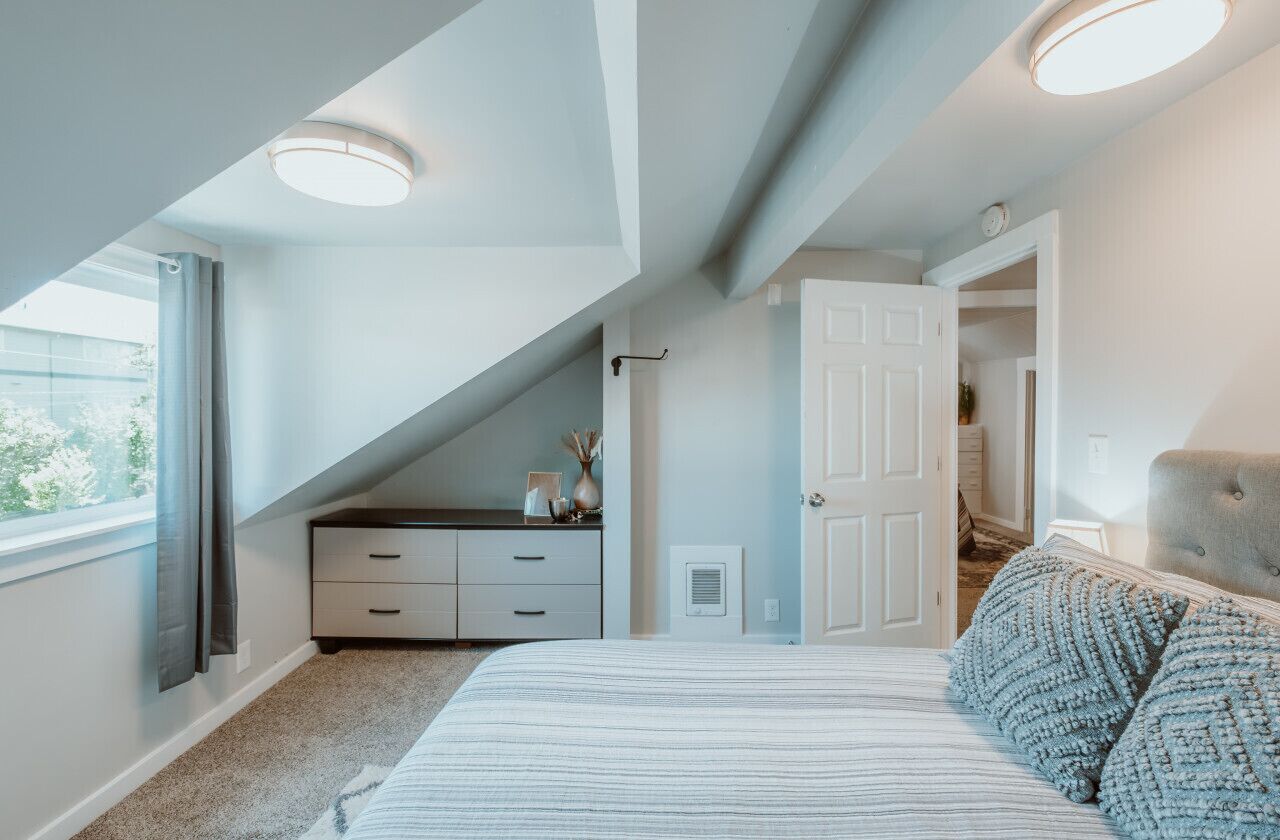 Quarto com teto inclinado, uma cama com colcha listrada, cômoda e uma cadeira.