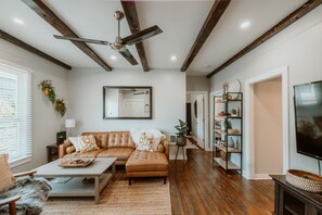 A living room with a brown leather sofa, a coffee table, and a television. There is a ceiling fan, wooden beams, and a bookshelf with decorative items. - A beautiful home near OSU campus (Corvallis)