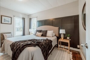 A bedroom with a large bed, a bedside table with a lamp, a framed picture on the wall, and a window with curtains. - A beautiful home near OSU campus (Corvallis)
