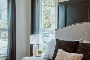 A bedroom with a large window, a bed with dark and light pillows, a bedside lamp, and a small plant. - A beautiful home near OSU campus (Corvallis)