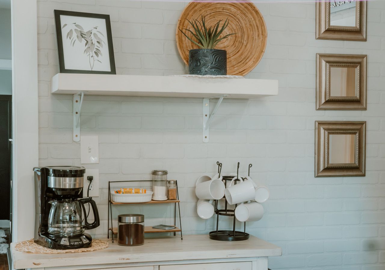A kitchen with a coffee maker, shelves with jars, a potted plant, and a mug rack.