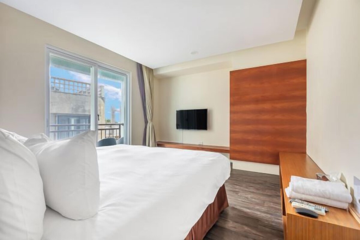 A hotel room with a large bed, a wooden headboard, a flat-screen TV, a bedside table with a remote control, and a window with a view of the city.