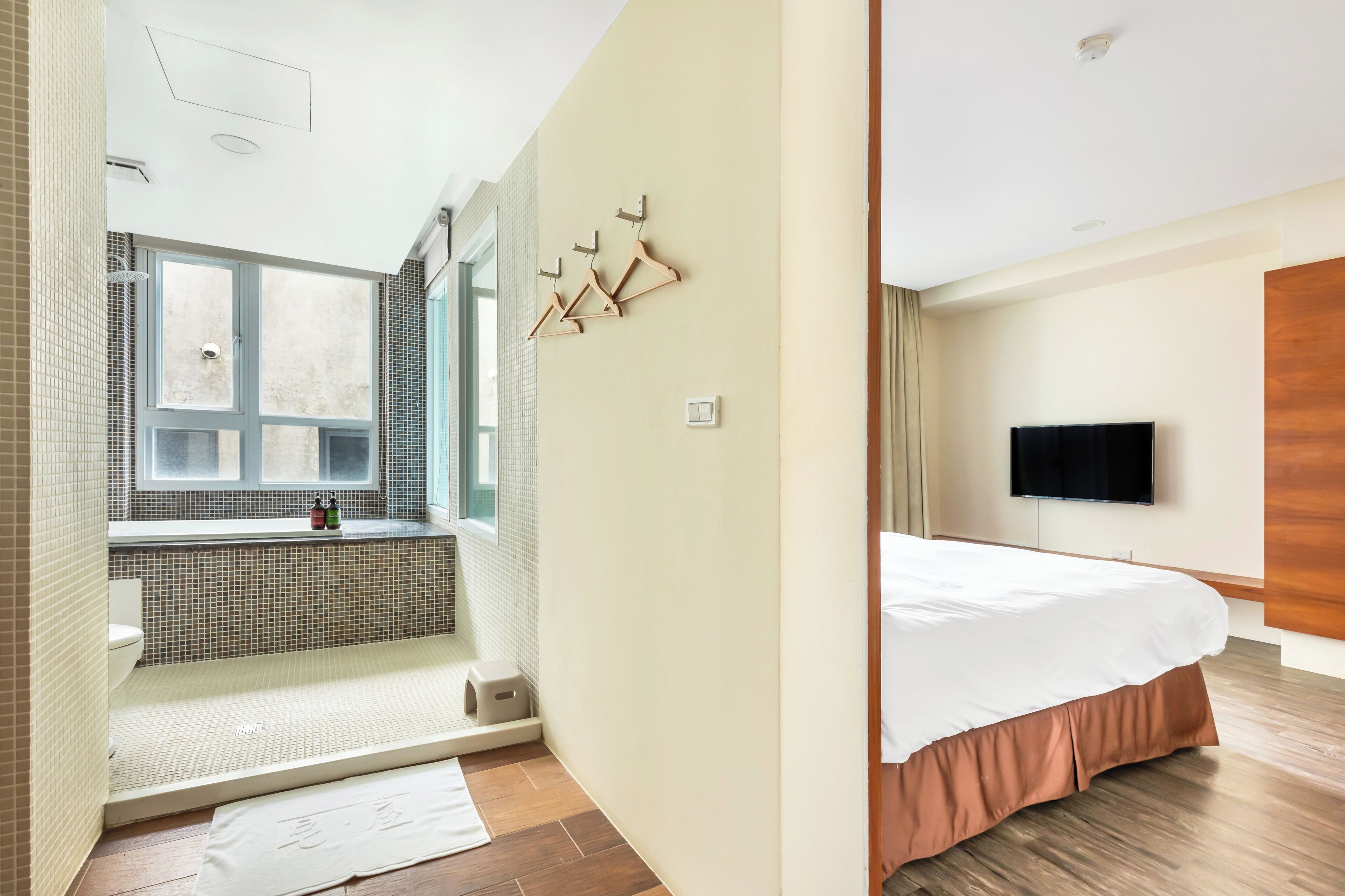 Une salle de bain moderne avec une baignoire, des toilettes et une fenêtre. On y trouve un lit en bois avec un matelas blanc et un couvre-lit marron. Un téléviseur à écran plat est fixé au mur.