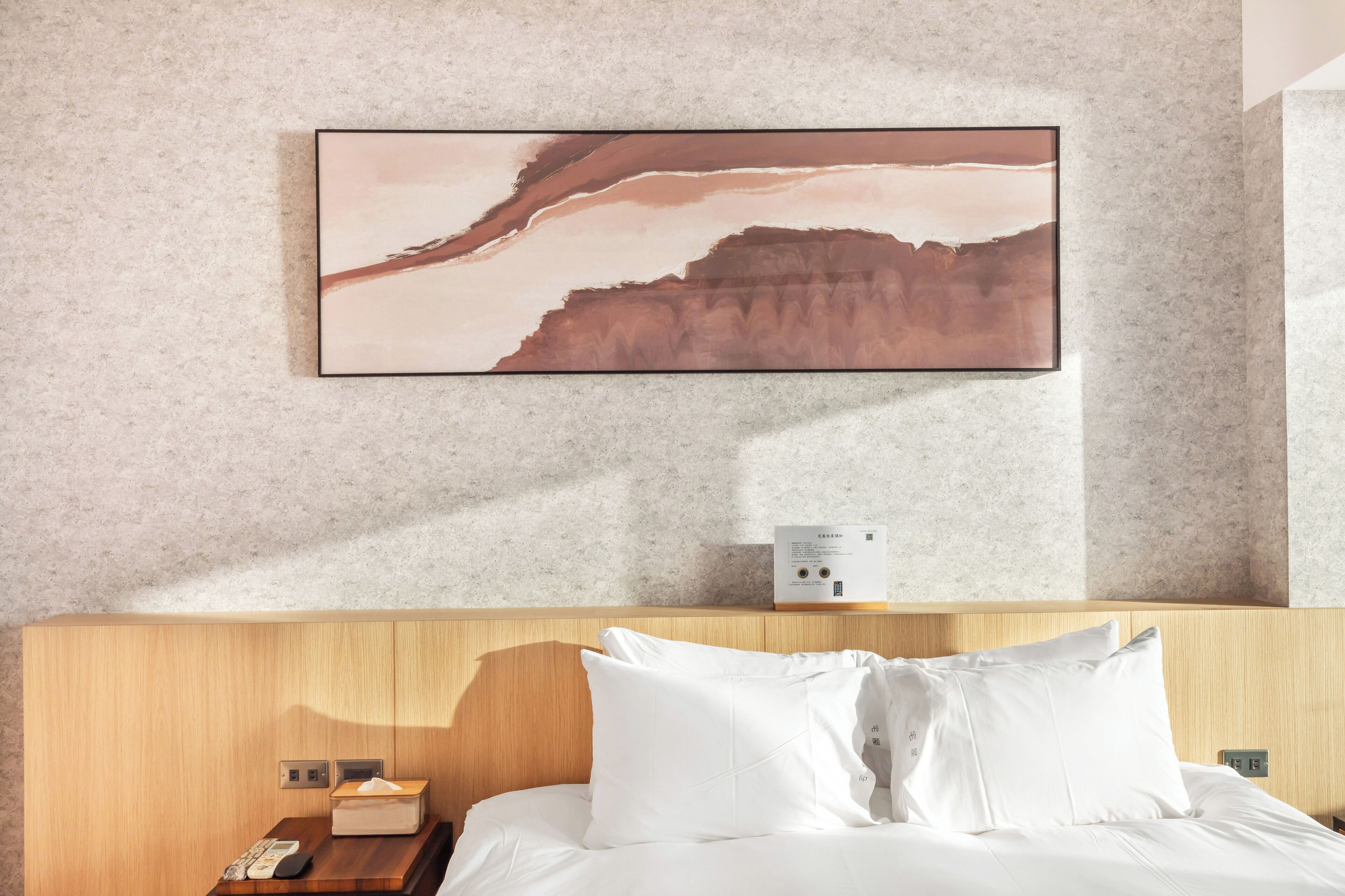 Ein Hotelzimmer mit einem hölzernen Kopfteil, einem Bett mit weißer Bettwäsche, einem Wandbild mit einer Landschaftsdarstellung sowie einem Nachttisch mit Taschentuchbox und Fernbedienung.