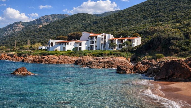 A coastal building with multiple balconies, situated on a rocky shoreline with clear blue waters.