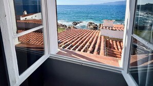 A room with a large window overlooking a coastal view, featuring a tiled roof and a clear sky.