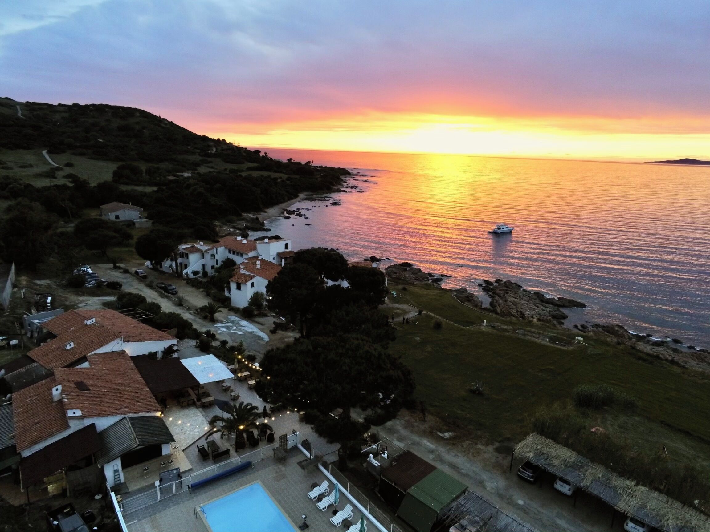 A coastal property with a swimming pool, a small boat docked near the water, and a view of the sunset over the sea.