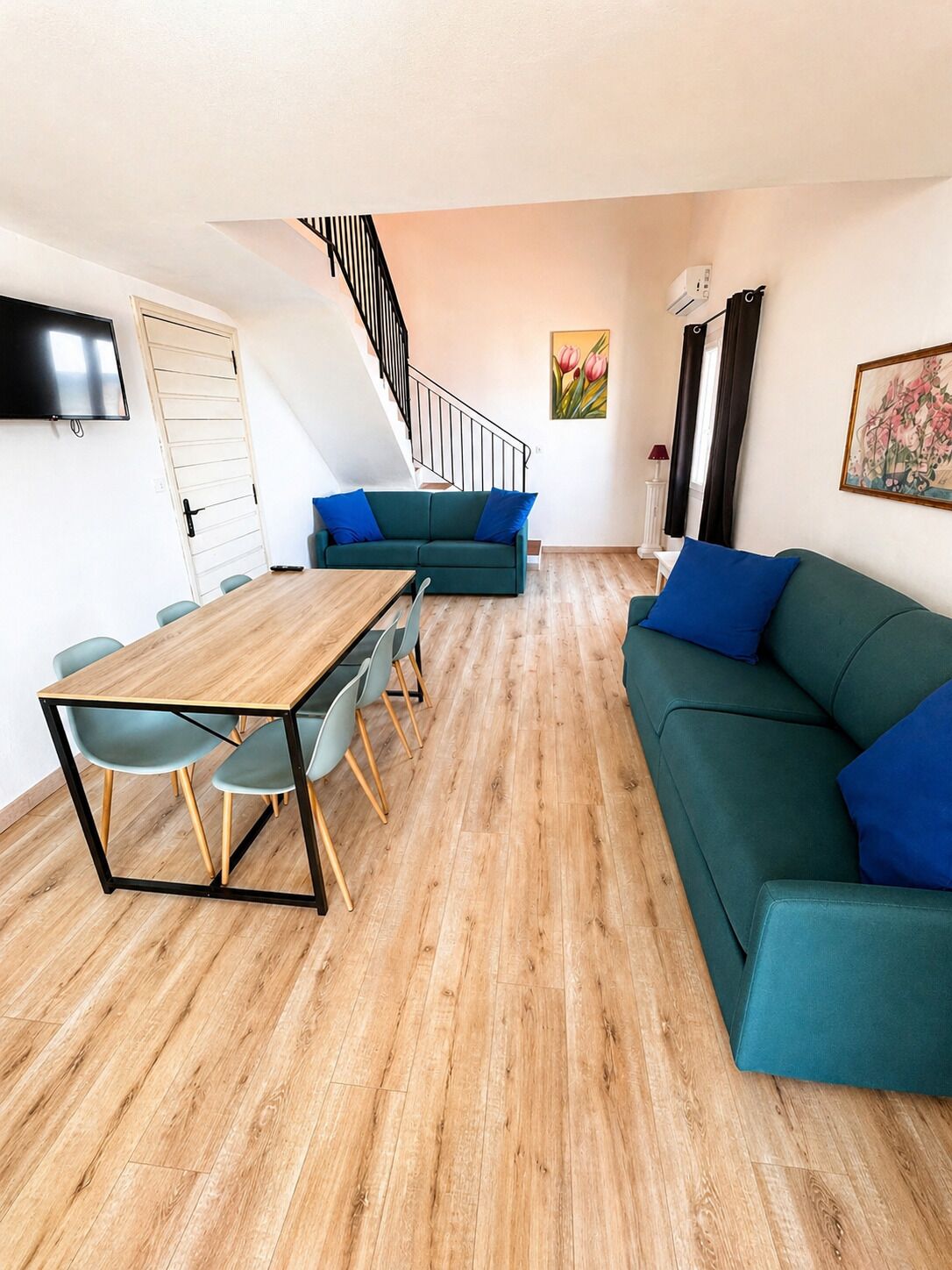 A modern living room with a wooden floor, a green sofa, a wooden dining table with chairs, a staircase, and a flat-screen TV mounted on the wall.