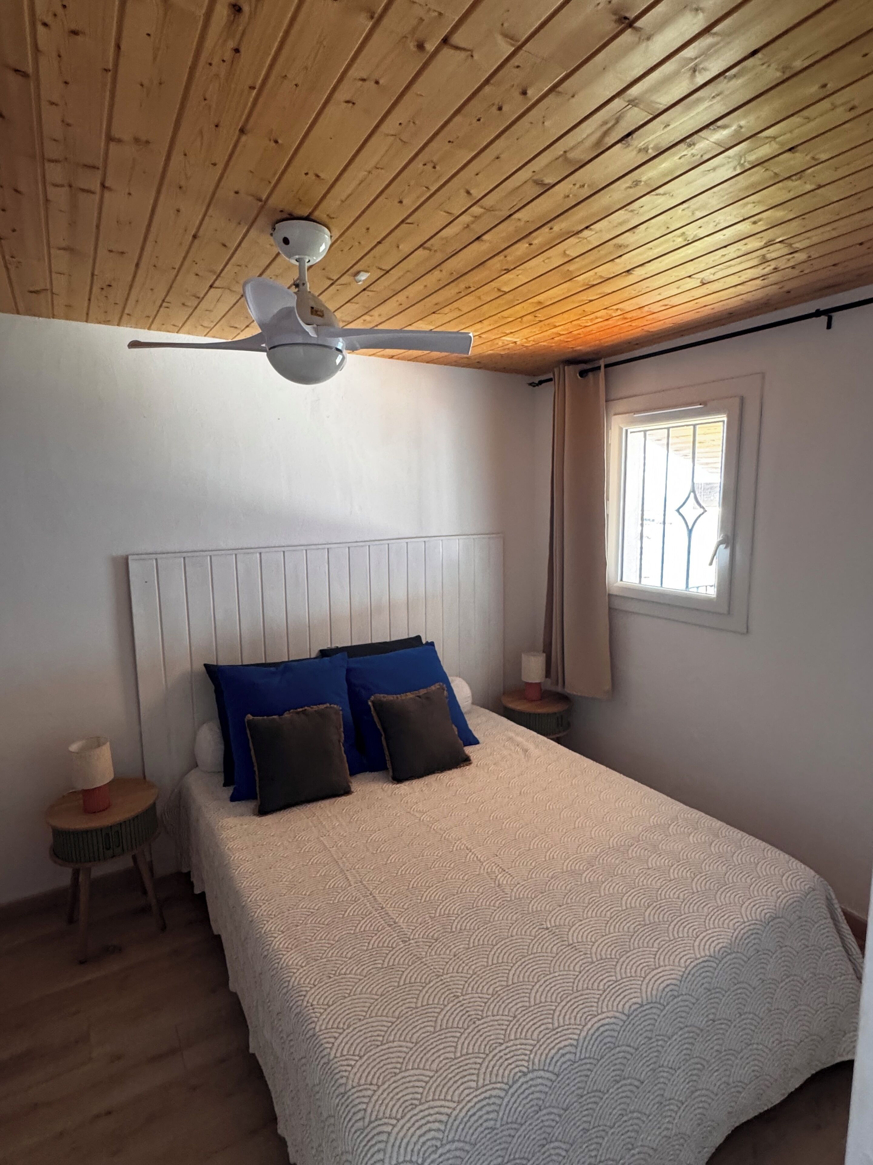 Een slaapkamer met een houten plafond, een bed met beddengoed met een patroon, een nachtkastje en een raam met gordijnen.