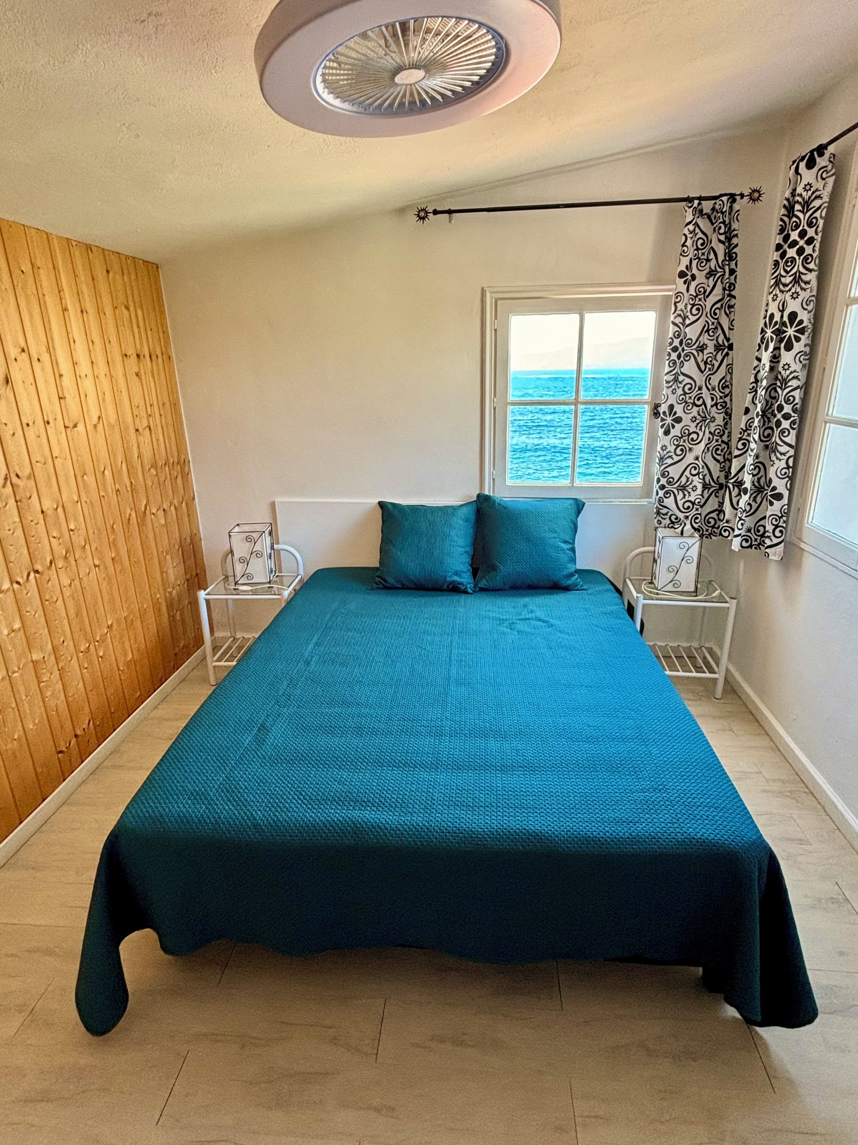 A bedroom with a large bed, a wooden wall, a window with a view of the sea, and a ceiling fan.