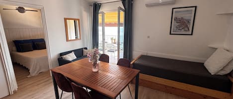 A room with a wooden dining table, chairs, a sofa, a bed, a mirror, and an air conditioning unit.
