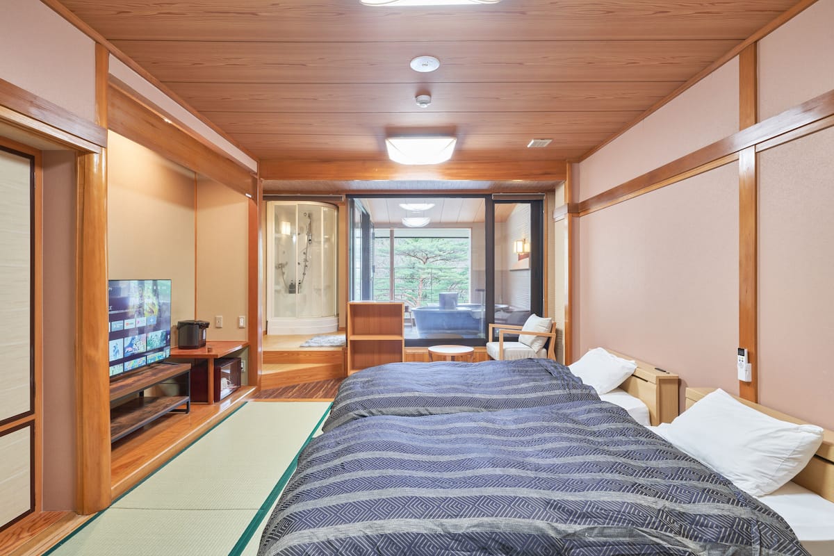 Una habitación tradicional japonesa con suelo de tatami, una cama grande con cubrecama estampado, un televisor sobre un mueble de madera y una puerta corredera que da a un balcón.