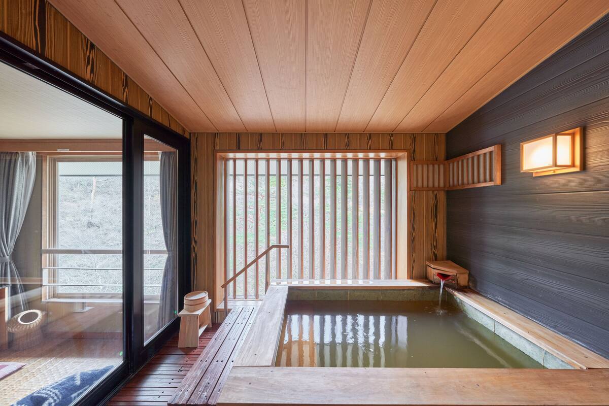 Un onsen japonés tradicional con suelos y bañera de madera, y vistas al exterior a través de amplios puertas correderas.