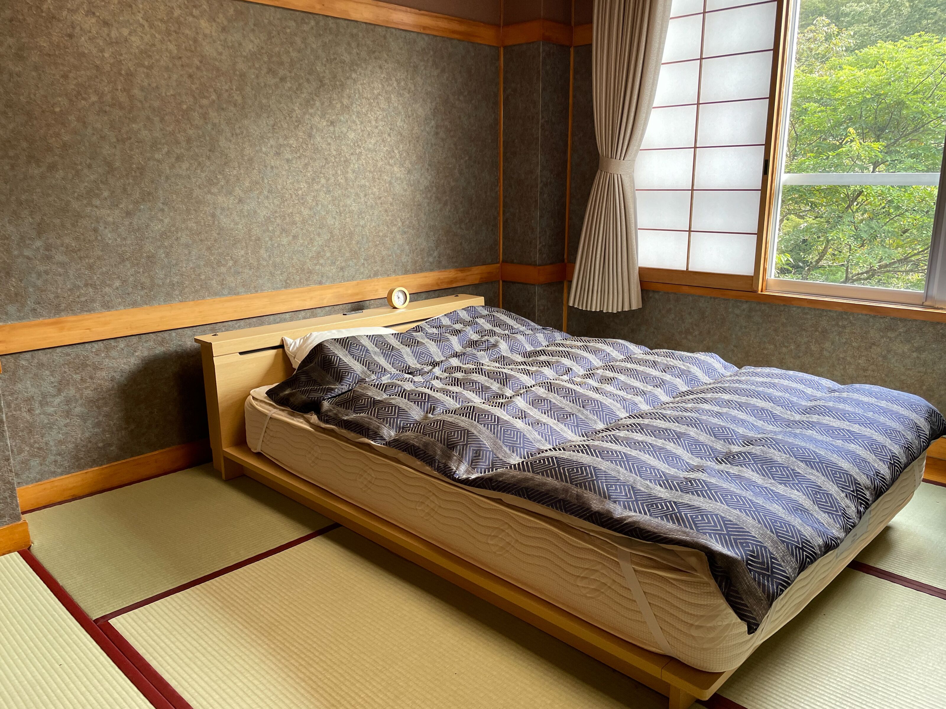 Um quarto tradicional japonês com tatames, cama de madeira baixa e janela com vista para vegetação.