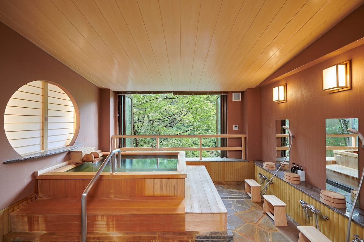 Un onsen japonés tradicional con bancos de madera, una fuente termal y vistas a árboles a través de amplios ventanales.
