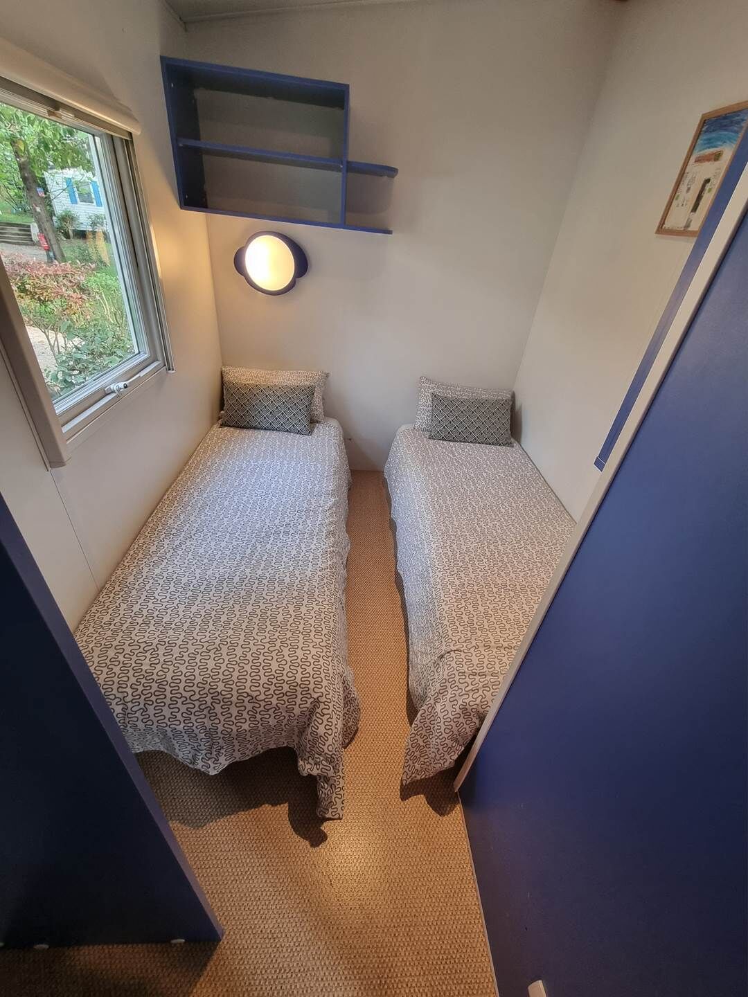 A small, narrow room with two beds, a window, and a wall-mounted shelf.