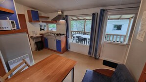 Une cuisine compacte équipée d’une cuisinière, d’un évier et d’un four à micro-ondes. Un coin repas avec une table et des bancs est aménagé. Une fenêtre avec des rideaux donne sur une terrasse extérieure avec des chaises.