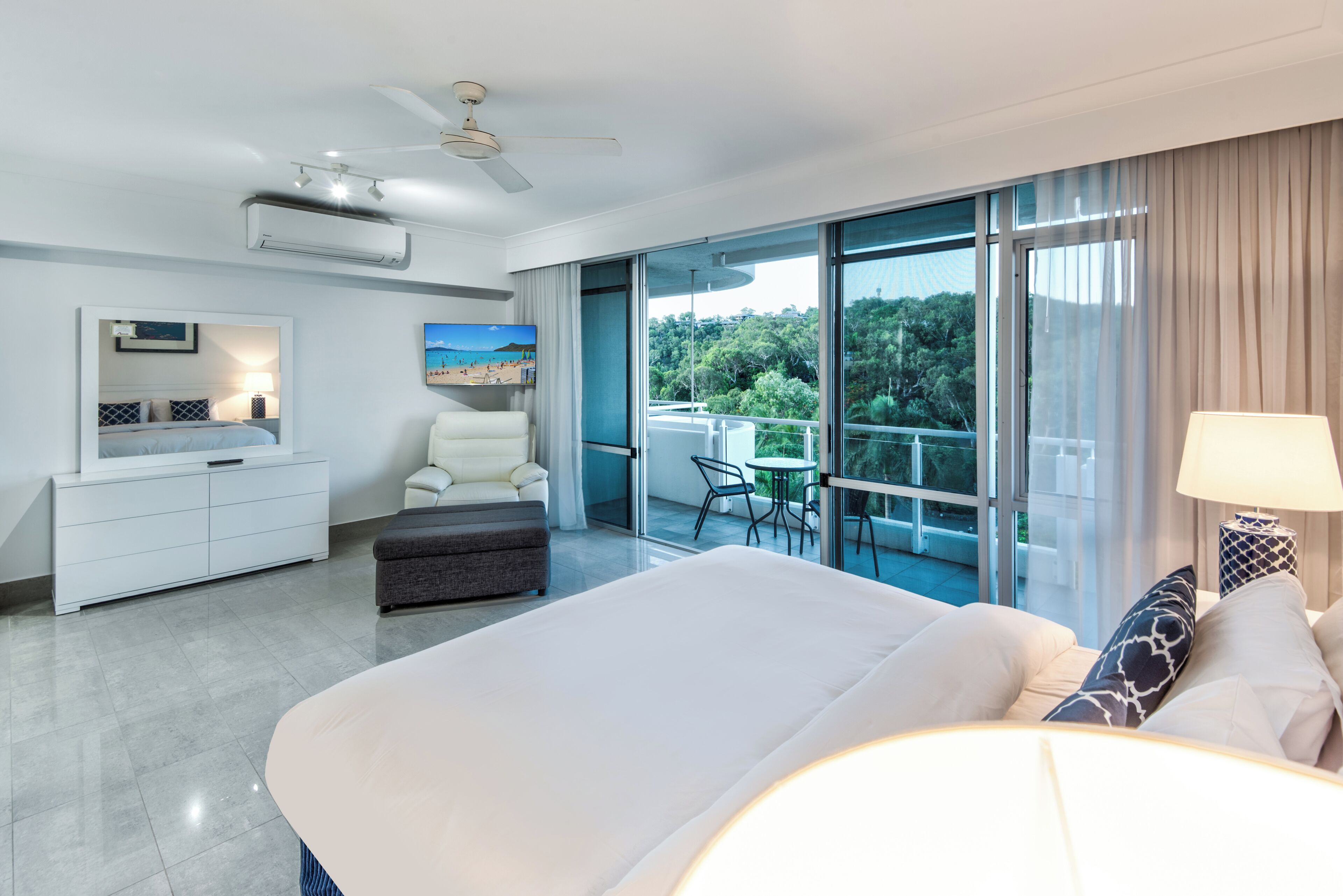 A modern hotel room with a large bed, a desk, a chair, a balcony with a view of trees, and a ceiling fan.