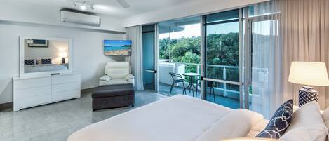 A modern hotel room with a large bed, a desk, a chair, a balcony with a view of trees, and a ceiling fan.