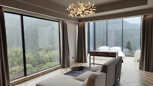 A modern hotel room with a large bed, a sofa, and a dining table. The room has large windows offering a view of a forested area. - Warm Place (Zhangjiajie)