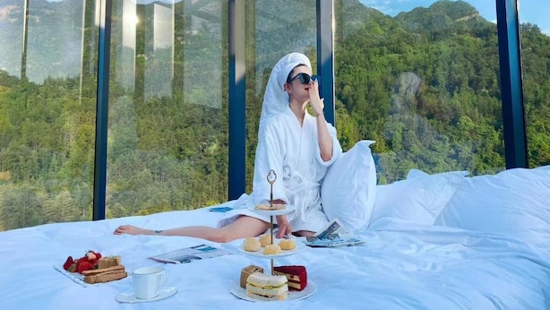 A person sitting on a bed with a scenic mountain view through large windows, enjoying a meal with sandwiches, pastries, and a cup. - Warm Place (Zhangjiajie)