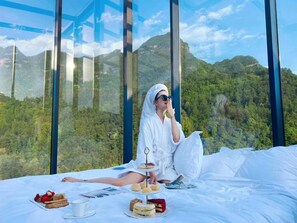 A person sitting on a bed with a scenic mountain view through large windows, enjoying a meal with sandwiches, pastries, and a cup. - Warm Place (Zhangjiajie)