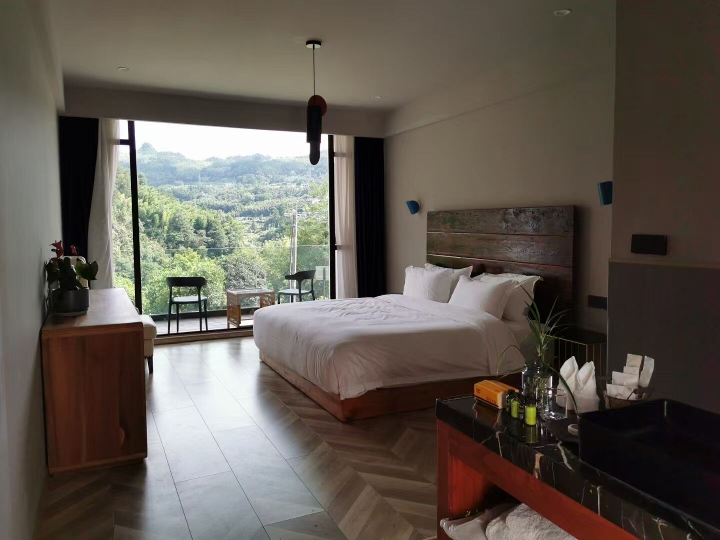 A modern bedroom with a large bed, a wooden nightstand, a TV, and a view of a lush landscape through large windows.