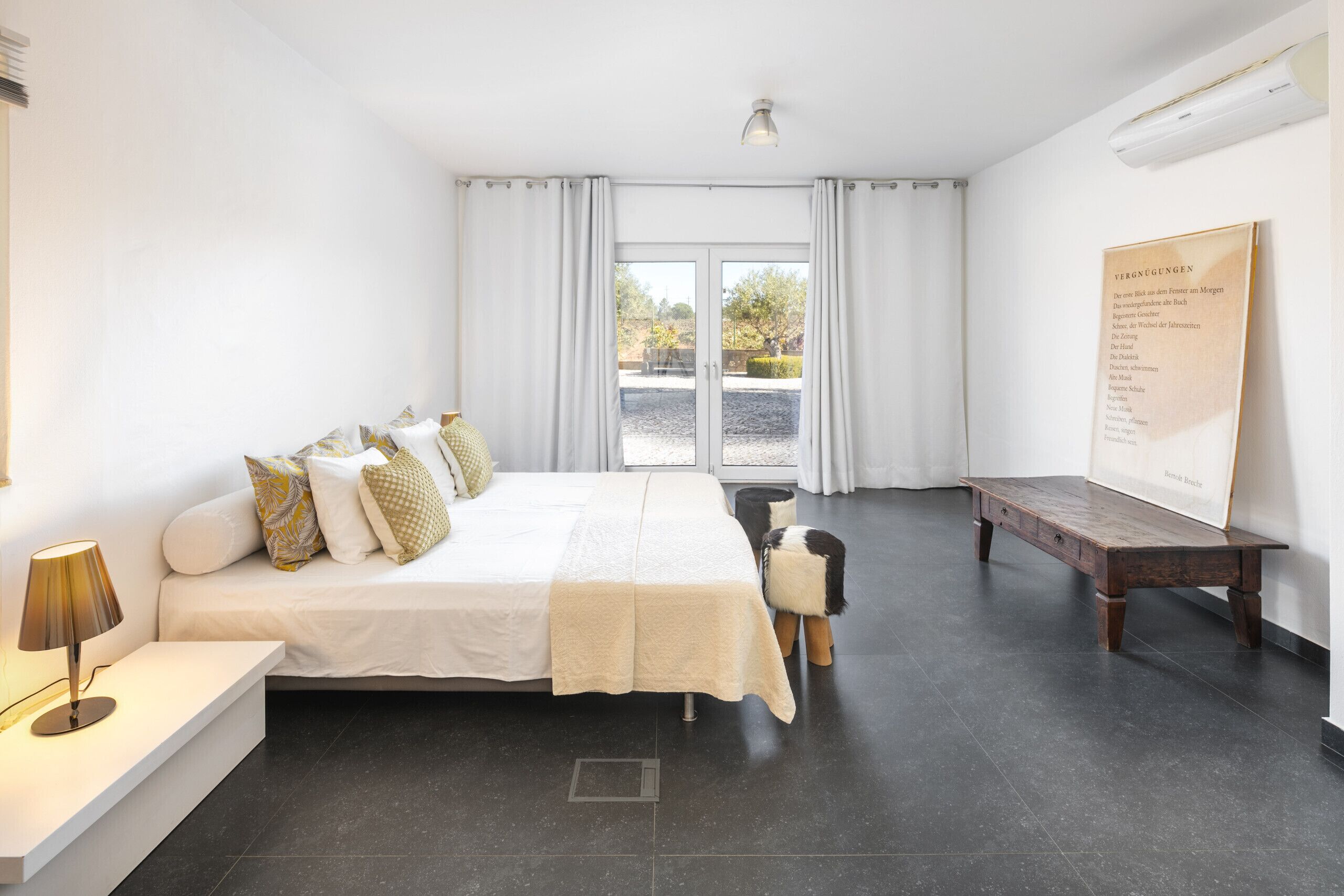 Ein modernes Schlafzimmer mit einem großen Bett, einem Nachttisch mit Lampe, einer hölzernen Bank und Blick ins Freie durch das Fenster.