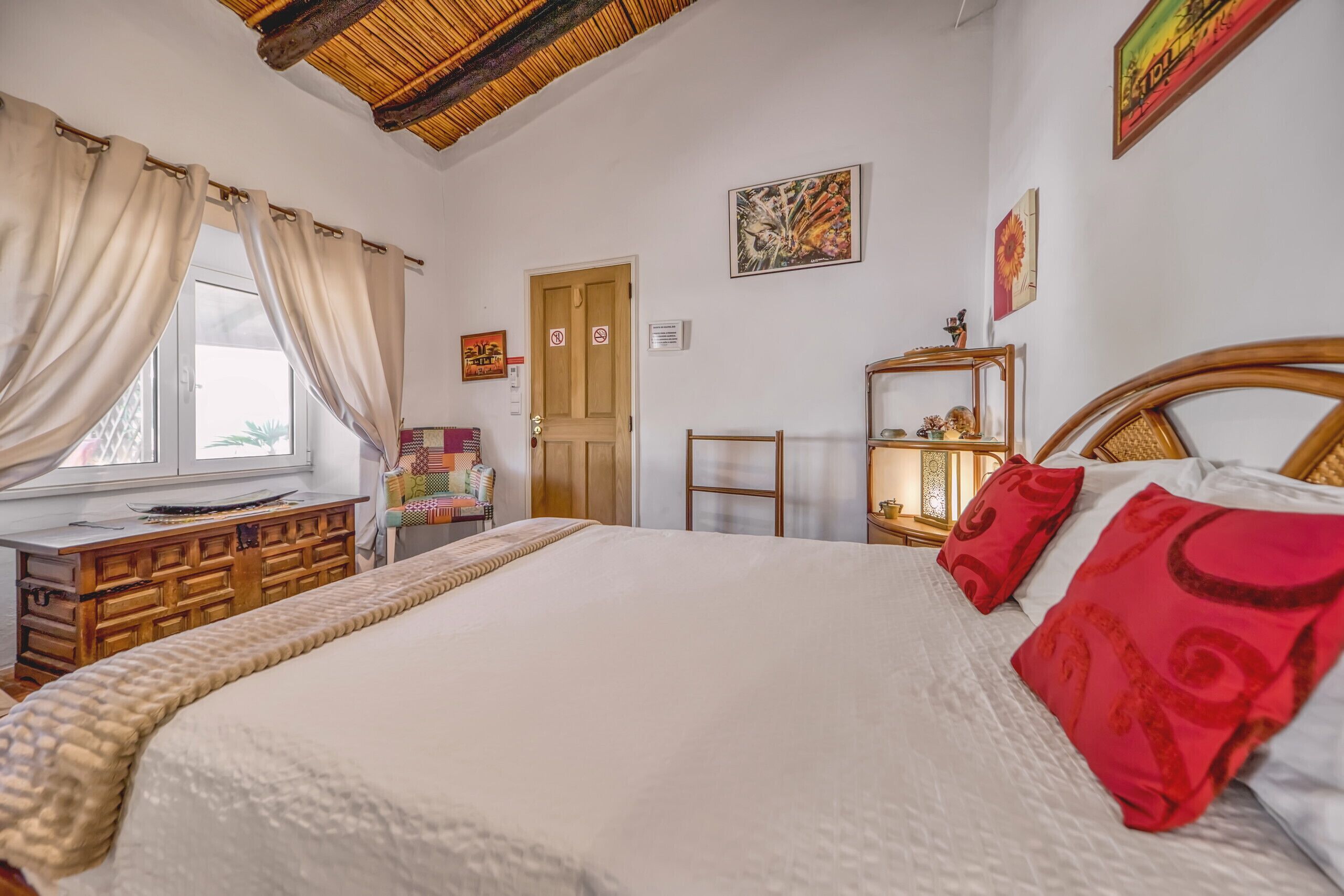 Une chambre à coucher avec un grand lit, une tête de lit en bois, une table de chevet, une commode, une fenêtre avec des rideaux et une porte.
