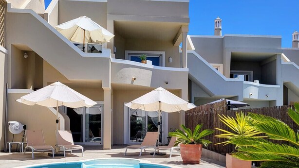 Une piscine entourée de chaises longues et de parasols, le tout bordé d’un bâtiment doté de balcons et agrémenté de plantes en pot.